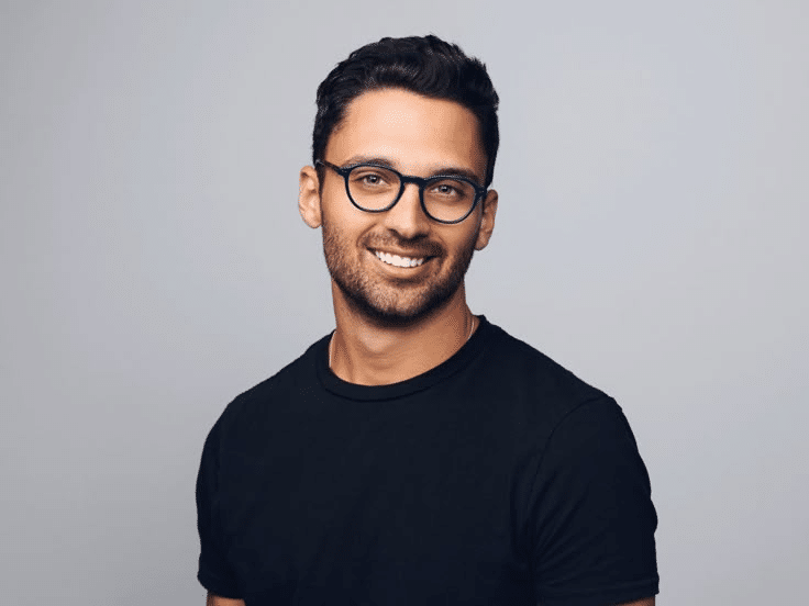 Muž s brýlemi v černém tričku stojí a usmívá se na kameru